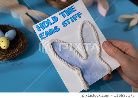 Person holding handmade greeting card with message HOLD THE EARS STIFF written in beautiful handwriting. Gesture shows excitement and joy for the upcoming event. Springtime holiday gift present zero Person holding handmade greeting card with message HOLD THE EARS STIFF written in beautiful handwriting. Gesture shows excitement and joy for the upcoming event. Springtime holiday gift present zero 136655173