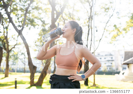 Pretty asian woman in sportswear standing with a hand on waist drinking water after exercise in park Pretty asian woman in sportswear standing with a hand on waist drinking water after exercise in park 136655177