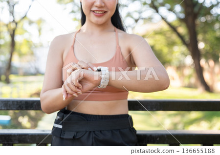 Close up sportswear woman looking at time on watch standing against railing after exercise in park Close up sportswear woman looking at time on watch standing against railing after exercise in park 136655188