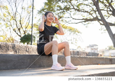 Asian woman in black sportswear wearing headphones sitting on concrete stairs after exercise in park 136655221