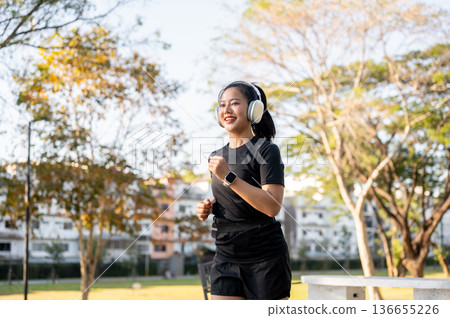Asian woman in sportswear listening to music in headphones while running jogging exercising in park. Asian woman in sportswear listening to music in headphones while running jogging exercising in park. 136655226