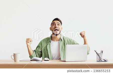 A man sits at a desk with a laptop and papers. He smiles and raises his arms in excitement. The setting is bright and simple. A mug and a pen holder are on the desk. 136655253