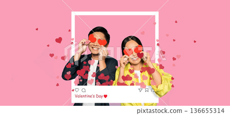 A couple is smiling and holding heart-shaped cutouts in front of a pink background. They are celebrating an event with a fun and joyful mood. Hearts float around them, adding to the excitement. A couple is smiling and holding heart-shaped cutouts in front of a pink background. They are celebrating an event with a fun and joyful mood. Hearts float around them, adding to the excitement. 136655314