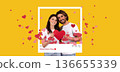 A couple stands together and smiles while holding red paper hearts. The background features a yellow color and floating heart shapes. This setup celebrates Valentine's Day in a fun way. 136655339