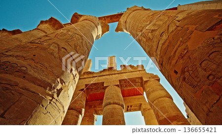 Ancient sandstone columns with carved hieroglyphs inside Karnak Temple complex in Luxor, Egypt, photographed from low angle against clear blue sky with dramatic sunlight Ancient sandstone columns with carved hieroglyphs inside Karnak Temple complex in Luxor, Egypt, photographed from low angle against clear blue sky with dramatic sunlight 136655421