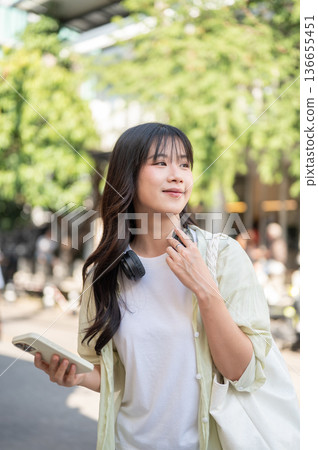 Pretty asian woman tourist holding phone looking away while standing or walking around local street Pretty asian woman tourist holding phone looking away while standing or walking around local street 136655451