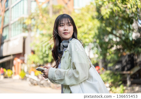 Pretty asian woman tourist holding phone carrying bag while standing or walking around local street. 136655453