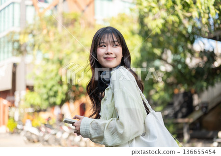 Smiling asian woman tourist holding phone carrying bag while standing or walking around local street Smiling asian woman tourist holding phone carrying bag while standing or walking around local street 136655454