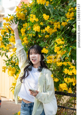 Pretty asian woman tourist holding phone raising one hand while standing under a yellow flower tree. Pretty asian woman tourist holding phone raising one hand while standing under a yellow flower tree. 136655457