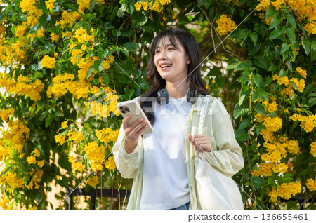 Smiling asian woman tourist holding phone and looking ahead while standing under yellow flower tree. Smiling asian woman tourist holding phone and looking ahead while standing under yellow flower tree. 136655461