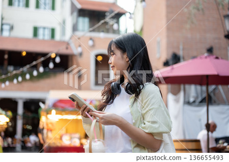 Asian woman tourist holding drink and phone while looking around walking in shopping mall or market. 136655493