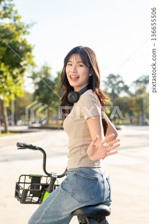 Happy asian woman waving a hand while riding green rental bicycle on pavement in park or city square 136655506