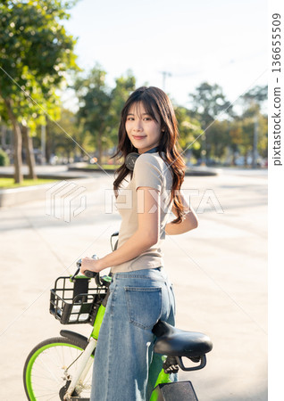 Pretty asian woman looking back as standing with green rental bicycle on pavement in park or square. 136655509