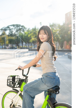 Pretty asian woman looking back while riding on green rental bicycle on pavement in park or square. 136655510