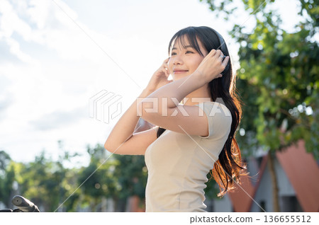 Pretty asian woman holding headphones listening to music while riding bicycle in park or city plaza. 136655512