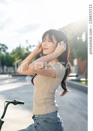 Pretty asian woman holding headphones while riding on bicycle in park or city plaza under sunlight. 136655515