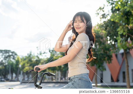 Pretty asian woman holding headphones while riding on a bicycle in park or city plaza under sunlight 136655516