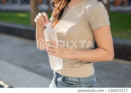 Close up of a woman holding a water bottle after drinking while standing in park or outdoors plaza. 136655517