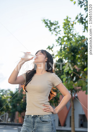 Pretty asian woman holding bottle drinking water while standing with hand on waist outdoors in park. 136655520