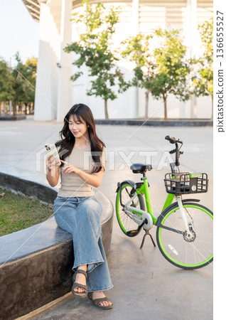 Pretty asian woman holding and looking at phone while sitting on bench aside bicycle in park plaza. 136655527
