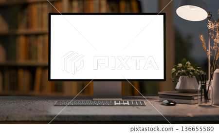 White screen computer with books and desk lamp on stone counter table and bookshelf in dim lit room. White screen computer with books and desk lamp on stone counter table and bookshelf in dim lit room. 136655580