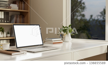 White screen laptop with books aside potted plant on counter table aside bookshelf and glass window. White screen laptop with books aside potted plant on counter table aside bookshelf and glass window. 136655589