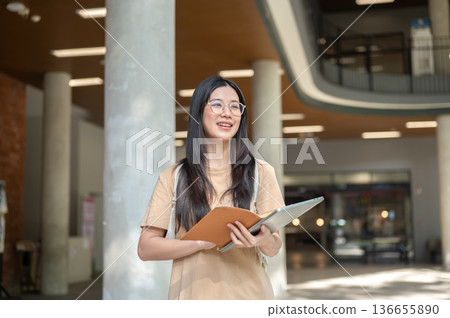 Glasses asian woman college student holding a book over laptop while standing or walking in building 136655890