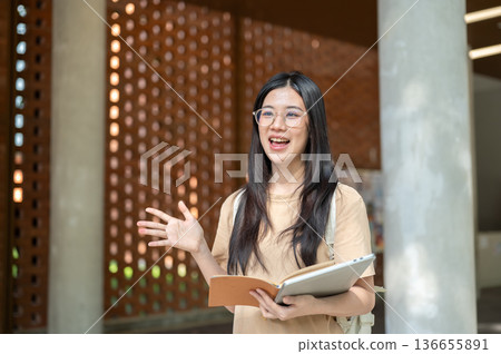 Glasses asian woman college student holding book and laptop waving a hand while walking in building. Glasses asian woman college student holding book and laptop waving a hand while walking in building. 136655891