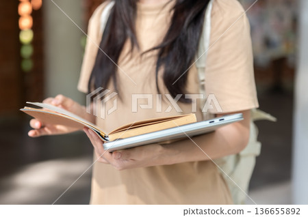 Close up of a woman college student reading a book over laptop while standing or walking in building Close up of a woman college student reading a book over laptop while standing or walking in building 136655892