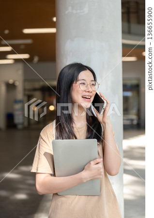 Glasses woman college student holding laptop talking on phone while standing or walking in building. 136655900