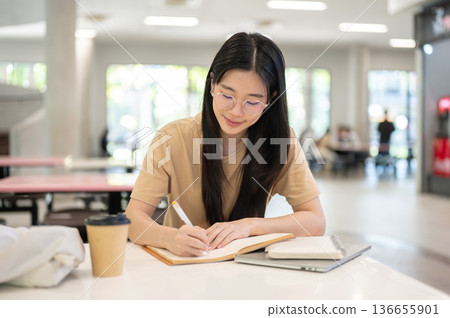 Glasses woman college student holding pen writing in notebook aside laptop at table in the building. Glasses woman college student holding pen writing in notebook aside laptop at table in the building. 136655901