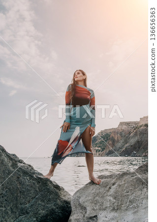 Woman Dress Rocks Beach - Fashion Model Posing on Rocks at the Beach in Colorful Dress. Woman Dress Rocks Beach - Fashion Model Posing on Rocks at the Beach in Colorful Dress. 136656363