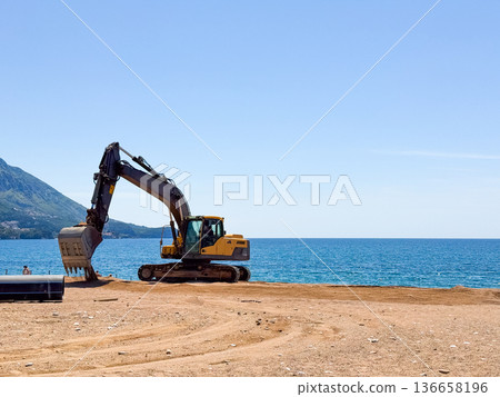 A yellow excavator standing on sandy beach near the sea and mountains. Industrial presence, construction and human activity by the coastline. A yellow excavator standing on sandy beach near the sea and mountains. Industrial presence, construction and human activity by the coastline. 136658196