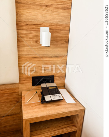 Bedside table with telephone, lamp, and notepad in hotel room. Communication, service, and assistance concept in hospitality environment for guest comfort. Bedside table with telephone, lamp, and notepad in hotel room. Communication, service, and assistance concept in hospitality environment for guest comfort. 136658625