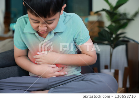 Asian boy holding belly suffering from severe abdominal pain with red inflamed area. Concept of stomach ache, indigestion, food poisoning, gastritis, and pediatric medical healthcare treatment. 136658710