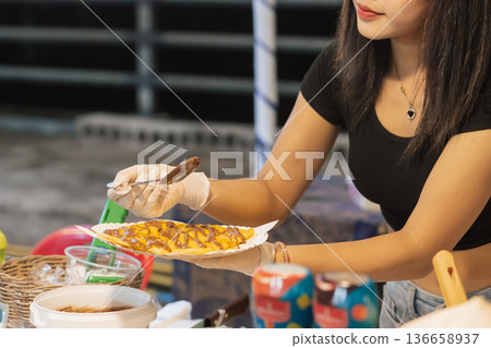A woman is making a sweet roti at a street stall in Bangkok. She is adding toppings with care while customers watch. This food is popular in Thailand and represents local street snacks. High quality A woman is making a sweet roti at a street stall in Bangkok. She is adding toppings with care while customers watch. This food is popular in Thailand and represents local street snacks. High quality 136658937