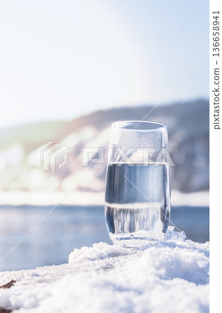 Water filled glass rests on fresh snow near a natural spring. Winter landscape surrounds the glass. Light reflects off the water. Nature looks bright and inviting in the cold weather. High quality Water filled glass rests on fresh snow near a natural spring. Winter landscape surrounds the glass. Light reflects off the water. Nature looks bright and inviting in the cold weather. High quality 136658941