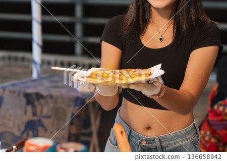 In a busy street food stall in Bangkok, a woman smiles as she holds a plate of sweet roti pancake. The warm lights create a lively atmosphere as customers enjoy traditional Thai snacks. High quality In a busy street food stall in Bangkok, a woman smiles as she holds a plate of sweet roti pancake. The warm lights create a lively atmosphere as customers enjoy traditional Thai snacks. High quality 136658942