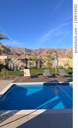 Afternoon Pool With Loungers And Umbrella, Clear Water Mirroring Mountain Ridge And Sky, Inviting Scene For Couples Getaway, Sunsetready Relaxation Or Boutique Hotel Leisure Moment 136659482