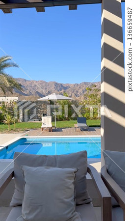Shaded Cabana Seating Overlooking Pool With Striped Pergola Shadows, Plush Cushions On Lounge Chairs And Distant Rocky Ridge Framing Calm Water For Cozy Respite Shaded Cabana Seating Overlooking Pool With Striped Pergola Shadows, Plush Cushions On Lounge Chairs And Distant Rocky Ridge Framing Calm Water For Cozy Respite 136659487