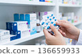 Person holding a pack of blue and white pills while standing in a pharmacy with various medications in view 136659933