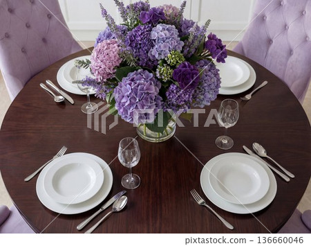 Large round table set with white plates and glassware, featuring a purple flower arrangement in a dining room 136660046