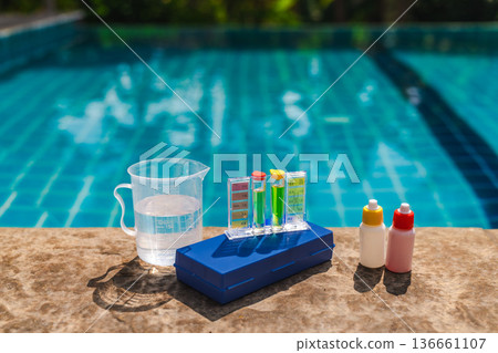 Pool water testing and treatment setup with pH and chlorine test kit, measuring cup and liquid chemicals on pool edge, outdoor swimming pool maintenance concept Pool water testing and treatment setup with pH and chlorine test kit, measuring cup and liquid chemicals on pool edge, outdoor swimming pool maintenance concept 136661107