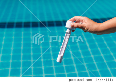 Hand holding pool thermometer measuring water temperature during swimming pool maintenance with clear blue tiles in background 136661114
