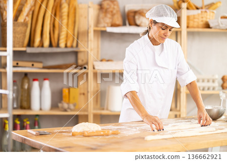 Female baker rolls dough into long sausages to be cut into pieces for baking buns Female baker rolls dough into long sausages to be cut into pieces for baking buns 136662921