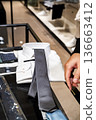 A customer compares two neckties on a glass counter inside a clothing store. The scene supports content about buying decisions and retail fashion choice. 136663412