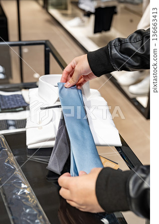 A customer compares two neckties on a glass counter inside a clothing store. The scene supports content about buying decisions and retail fashion choice. 136663413
