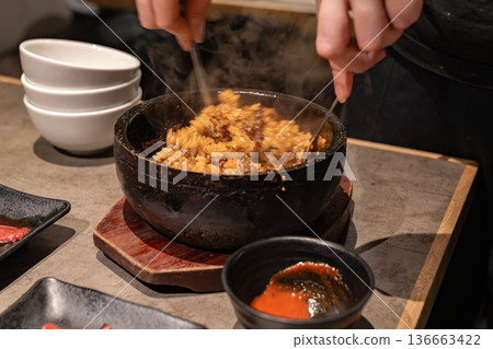Hands mixing cooked rice inside a hot stone bowl with visible steam. The action shows active food preparation at the table. 136663422