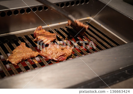 Beef slices cook on a metal grill with visible heat and browning surface, reflecting a professional restaurant environment and active food preparation process. 136663424