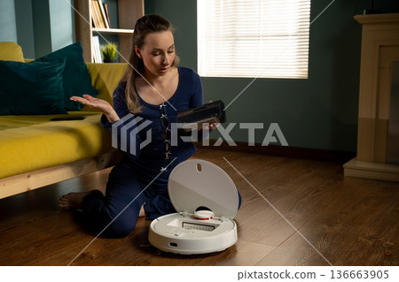 Woman sitting on a wooden floor, holding a dirty dustbin and looking confused about how to clean or maintain her automatic robotic vacuum cleaner in a modern living room Woman sitting on a wooden floor, holding a dirty dustbin and looking confused about how to clean or maintain her automatic robotic vacuum cleaner in a modern living room 136663905